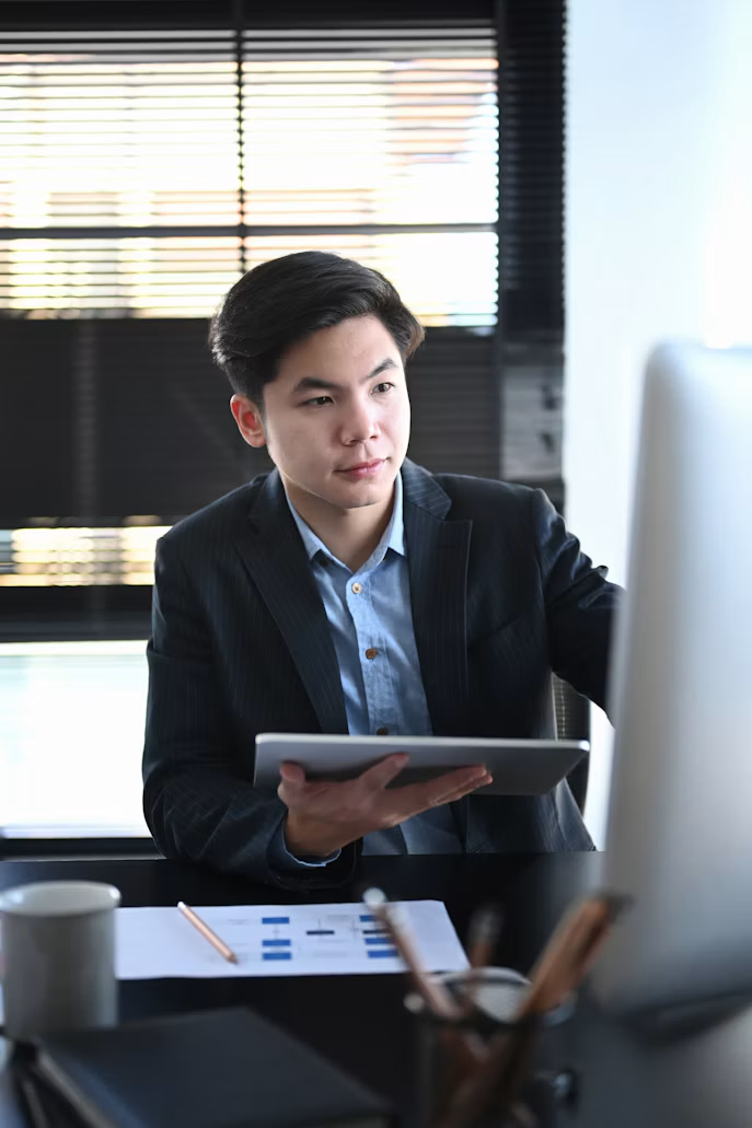 A consultant in a dark blazer reading a tablet at a desk in front of office blinds