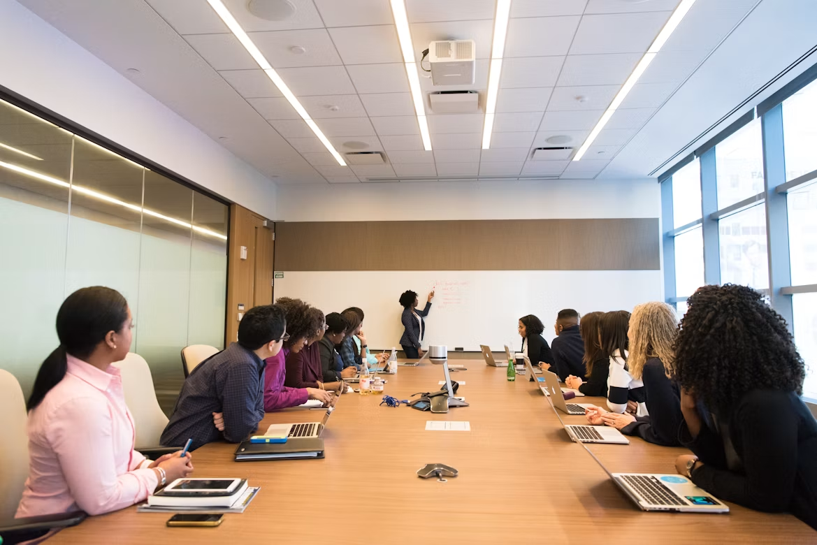 Colleagues seated the length of a long boardroom table while one of them presents at the whiteboard