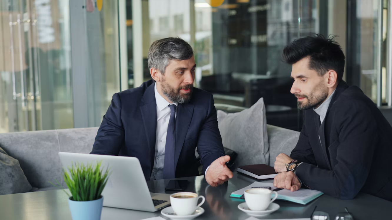 Two executives in suits talking across a meeting table in a glass-walled office