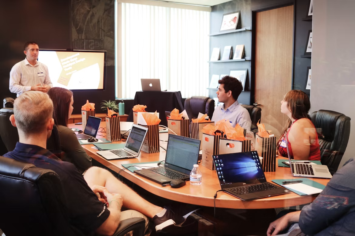 A trainer standing beside a presentation screen while a seated group follows along on laptops